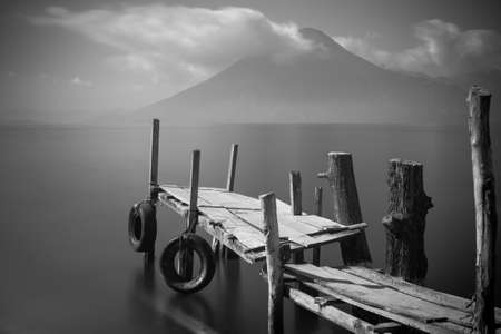 Old rickety wooden pier juts out into lake Atitlan  Guatemala  with volcano in background   Black and Whiteの写真素材