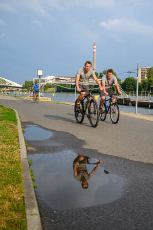 PRAGUE - JULY 20, 2019: Cyclists on a riverside path with reflection in water puddleのeditorial素材