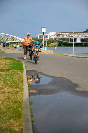 PRAGUE - JULY 20, 2019: Young child on a bike reflected in a puddle of water and followed by his mother pushing a bicycleのeditorial素材