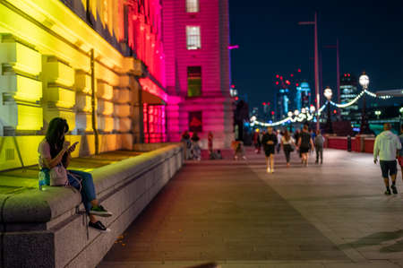 LONDON - SEPTEMBER 14, 2021: A young woman uses a smartphone while sat on a wall next to colorfully lit County Hall at nightのeditorial素材