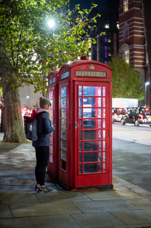 LONDON - NOVEMBER 2, 2021: A young woman with backpack outside of traditional British Red Telephone Boxes at nightのeditorial素材