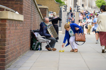 LONDON - May 21, 2022: Woman gives money to Elderly busker with white beard and accordion. Tourists...