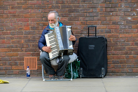 LONDON - May 21, 2022: Elderly busker with white beard plays accordion next to old red brick wallのeditorial素材