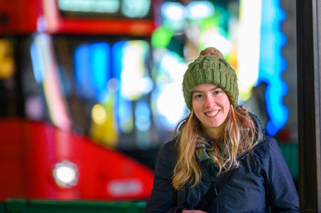 Smiling blonde female in bobble hat with Red London Double Decker bus and neon signs out of focus in the backgroundの写真素材
