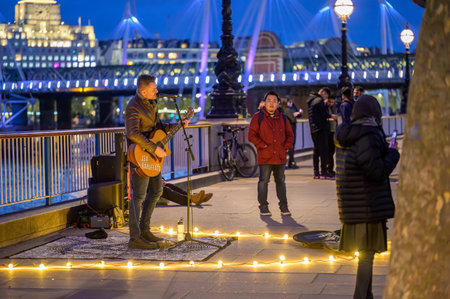 LONDON - April 22, 2023: Night entertainment: Busker enchants tourists on Southbank, London, under warm lighting against a deep blue evening sky.のeditorial素材