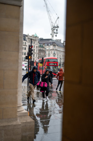 LONDON - April 24, 2023: Caught in the downpour, people dash for shelter on a bustling London street. A red double-decker bus looms in the background, framed by stone pillars.のeditorial素材