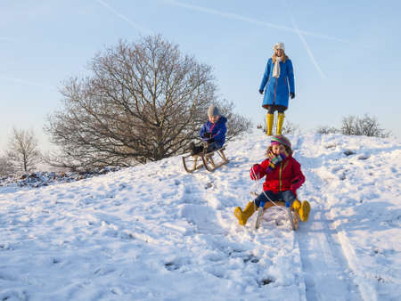 small boy and girl sledging down a snow covered dune while mother watchesの写真素材