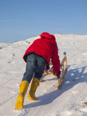 small girl in colourful winter clothing pushing a wooden sledge to the top of the hill in a snow covered dune landscapeの写真素材