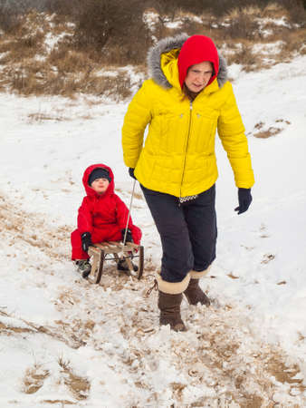 smiling mother in colourful winter clothes pulls small boy in red winter suit on a wooden sledge through snow covered dunesの写真素材