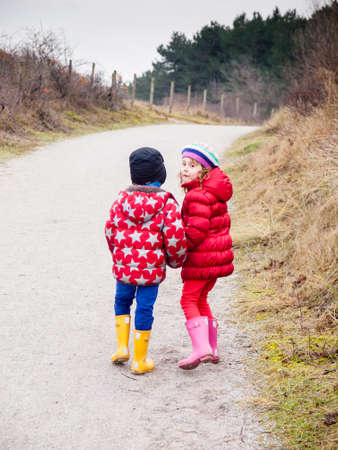 small boy and girl in winter clothing walking hand in hand along a path througha wooded landscapeの写真素材