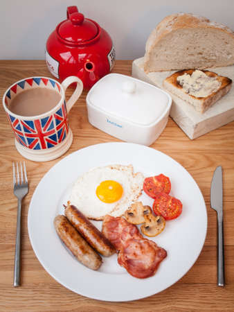 English breakfast opf bacon eggs and sausage on a wooden table with teapot, cup pf tea, butter dish and toast の写真素材