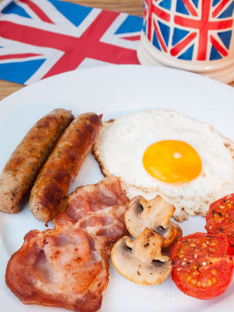 Close-up of cooked English Breakfast on a wooden table with cup of tea  in union jack mug and british flag behind in portraitの写真素材