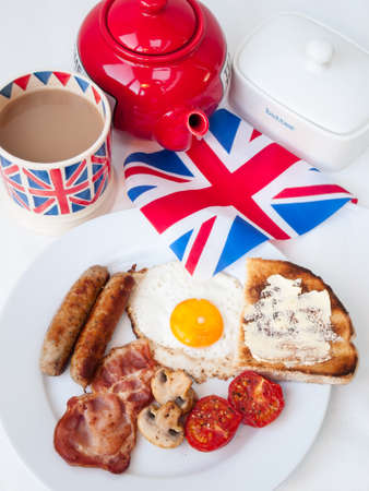 English fried breakfast on a white table top with cup of tea in union jack mug, red teapot, buttered toast  and british flag behindの写真素材