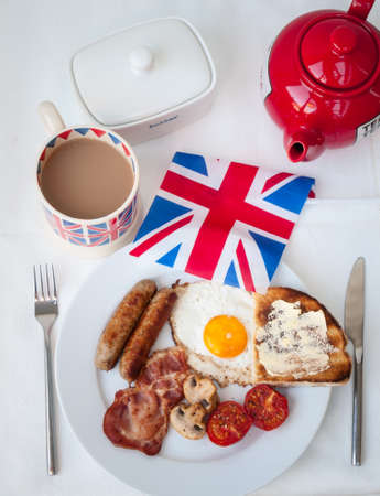 English fried breakfast on a white table top with cup of tea in union jack mug, buttered toast , butter dish , red teapot and british flagの写真素材