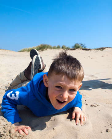 small smiling boy lying on sand at the base of a  sand dune on a bright sunny spring day under a blue skyの写真素材
