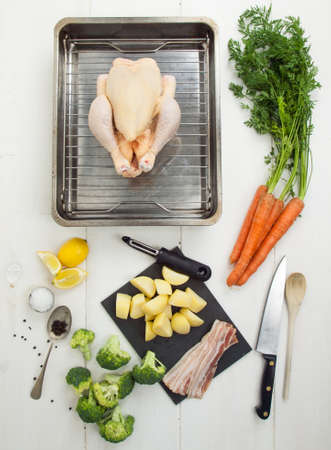 ingredients for a roast chicken dinner with whole chicken, vegetables and utensils on a rustic white table topの写真素材