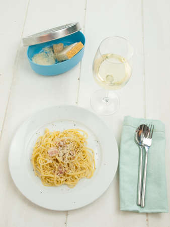 plate of spaghetti carbonara in a rustic table setting with grated cheese, fork and spoon, and a glass of white wineの写真素材