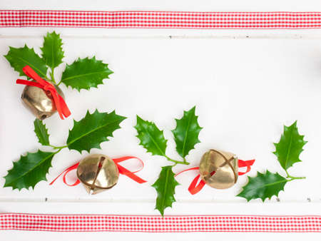 christmas sleigh bells and green holly leaves with checked red ribbon against a rustic white table topの写真素材