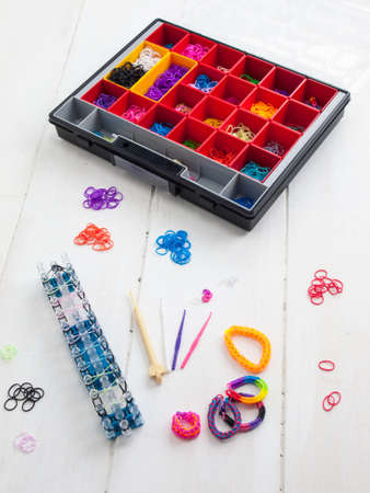 colourful elastic loom bands with band loom , bracelets, tools and hobby box against a white table...