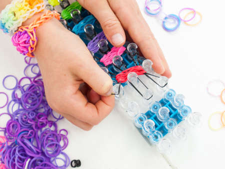 closeup of childs hands making a braclet with colourful elastic loom bands on a band loom against a white table topの写真素材