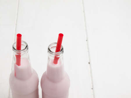 detail of two bottles of pink milkshake with red straws against a white wooden table topの写真素材