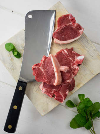 overhead view of uncooked lamb chops on a chopping board with a meat cleaver and and fresh mint leaves with rustic white table behindの写真素材