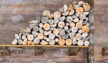stack of sawn logs against and olf cabin wall with falling snow in teh foregroundの写真素材