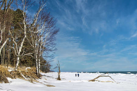 Beneath blue skies and white clouds on a winter's day, a couple walks along the frozen Lake Michigan shoreline at Harrington Beach, near Lake Church, WI.の写真素材