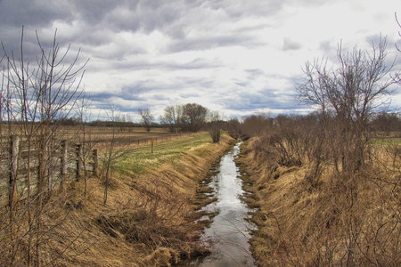 Near Oshkosh, Wisconsin, the Wiouwash State Trail runs alongside a steam of water, surrounded by leafless trees and dry grasses under a cloudy sky.の写真素材