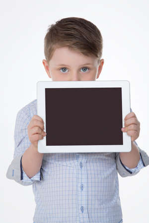 shy male kid hides his mouth behind white tabletの写真素材