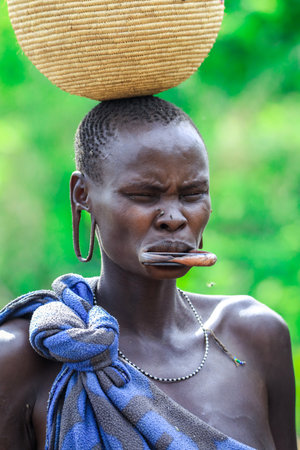Omo River Valley, Ethiopia - November 29, 2020: Portrait of African Woman with a big traditional wooden plate in the lower lip and Wicker basket on the head in the local Mursi tribe villageのeditorial素材
