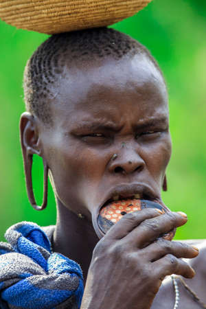 Omo River Valley, Ethiopia - November 29, 2020: Portrait of African Woman with a big traditional wooden plate in the lower lip and Wicker basket on the head in the local Mursi tribe villageのeditorial素材