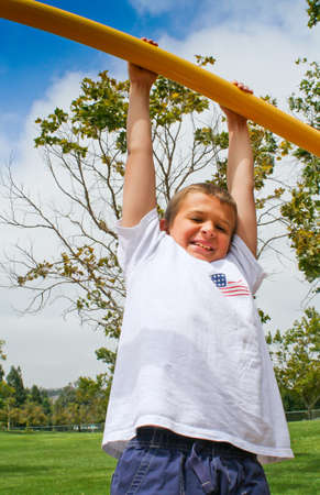 Happy youth hanging from high bar in park against blue skyの写真素材