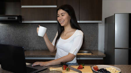 young woman in the kitchen looking at the laptop while eating a healthy breakfast