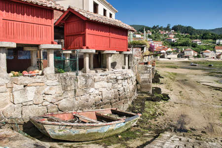 Old abandoned rowing boat next to traditional red granaries at low tide in the town of Combarro,...