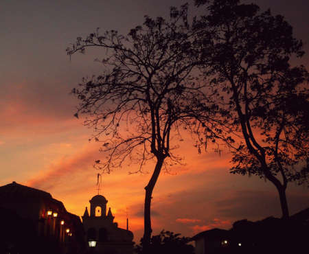 Red sunset with the silhouette of a colonial town, leafy trees and the belfry of a churchの写真素材
