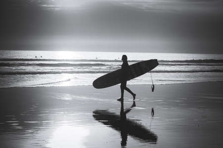 BOY WALKING WITH HIS SURFBOARD BY THE BEACH AT SUNSETの写真素材