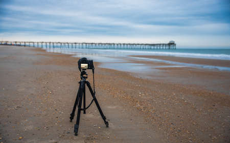 Camera taking photo of the sunrise with the Atlantic Beach pier in the background with sand, ocean, waves and blue cloudsの写真素材