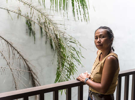 Asian Woman standing on balcony with white wall and tree behind her