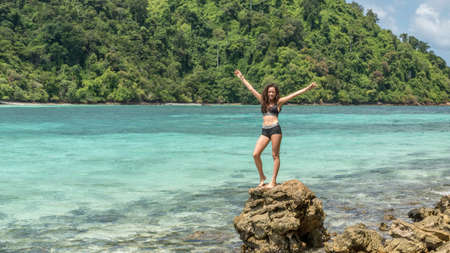 Young teen girl in black bathing suit on tropical island standing on rock with arms upの写真素材