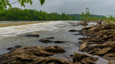 Woman in pink sweater sitting by the side of the river looking at the waterfallの写真素材