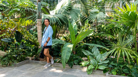 Asian woman in sneakers and skirt leaning on wall in botanical garden surrounded by plants