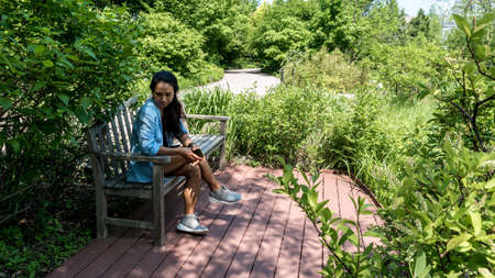 Asian woman in skirt sitting on bench on brick patio surrounded by plantsの写真素材