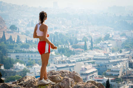 Teen in red shorts and white sneakers standing on rock on mountain with city of Athens in the backgroundの写真素材
