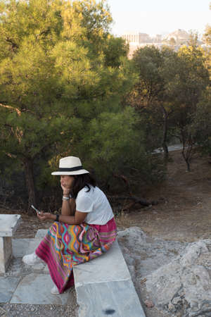 Asian woman in white t-shirt in Athens in white hat, sitting on marble bench in garden on Phillippas Hillの写真素材
