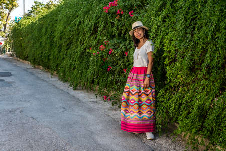 Asian woman in white t-shirt in Athens in white hat and sarong standing in front of ivy wall with flowers on small roadの写真素材