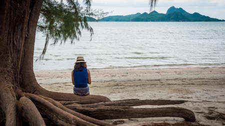 Woman sitting on large tree roots on the beach looking at her phone in Khoa Lommuak thailand