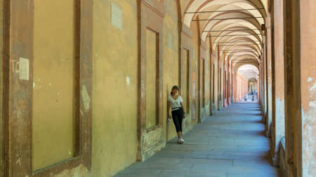 Asian woman in hat, tshirt and sneakers leaning against wall of portico in Bologna, Italy