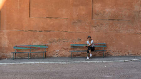 Woman in hat, tshirt, black pants and sneakers sitting on park bench with legs crossed in Bologna...