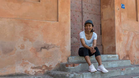 Woman sitting on steps in front of doorway with cap and sneakers in Bologna Italy looking at photographerの写真素材